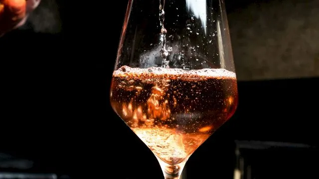 A bartender pours amber wine into a stemmed glass, sparkling as it fills under moody bar lighting.