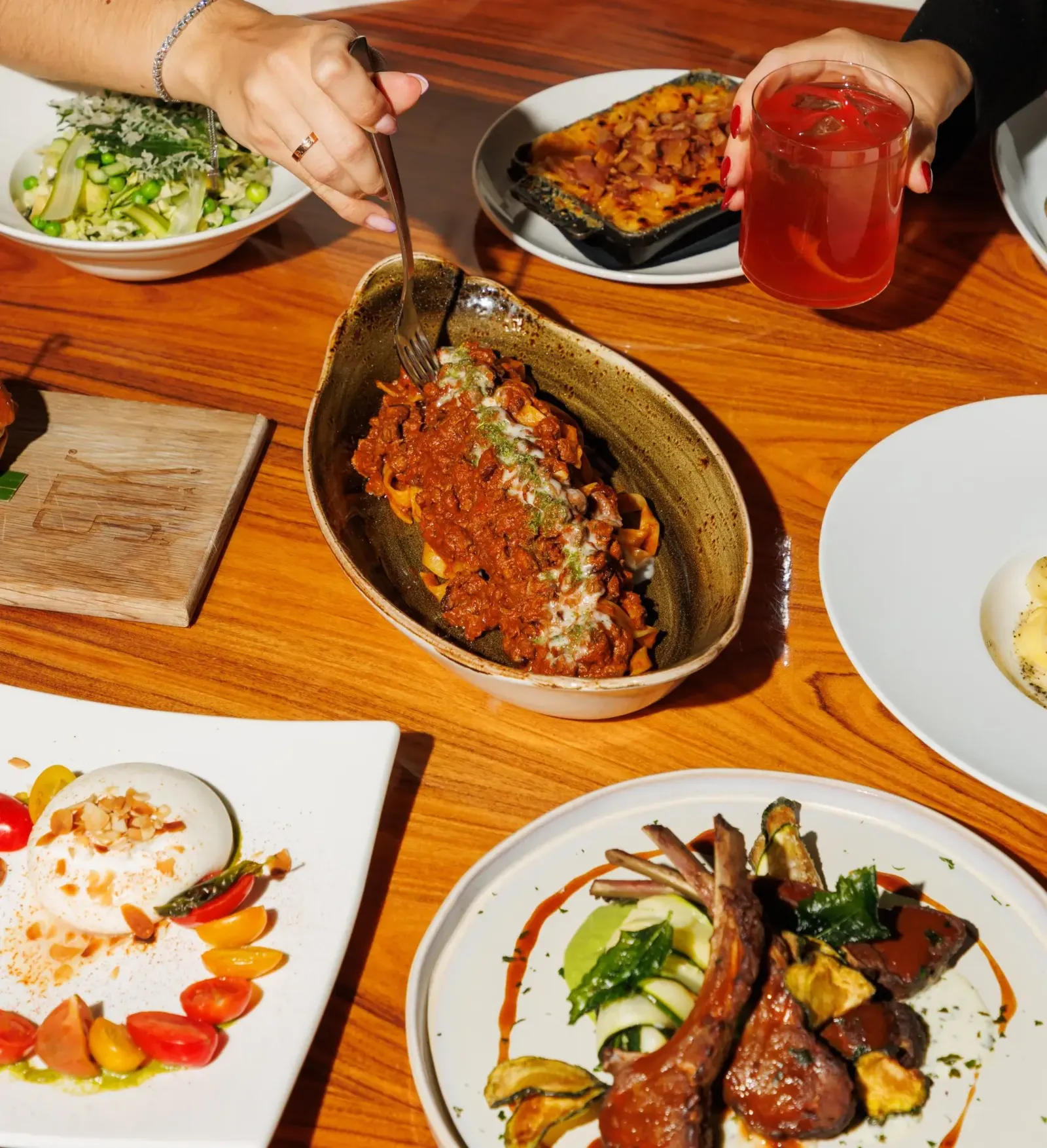 Overhead view of a shared table spread with pasta in meat sauce, lamb chops, burrata salad, side dishes, and hands reaching in with a fork and a red cocktail.