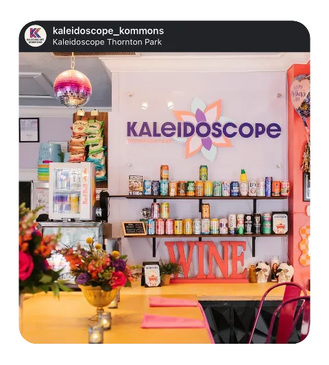 Interior of Kaleidoscope Thornton Park with a wall decorated with a large sign that says 'Kaleidoscope' with a flower motif, shelves filled with beverages, and a table with a flower arrangement in the