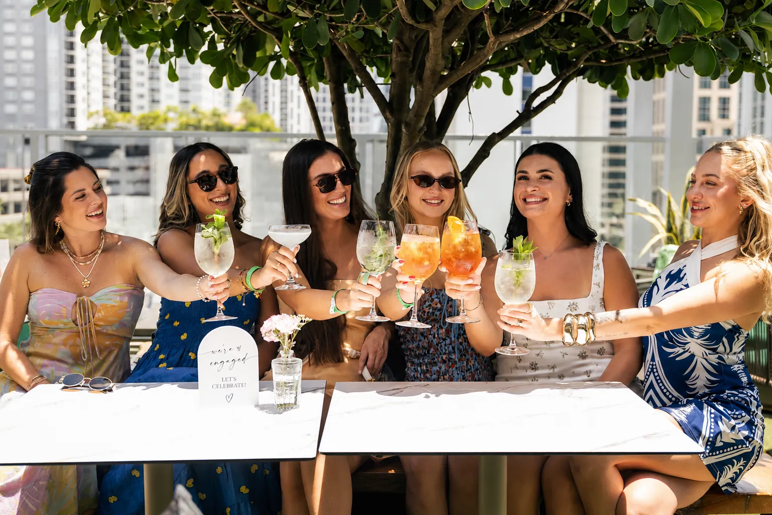 Six women in summer dresses raise colorful cocktails in a rooftop toast, smiling under a tree with city buildings in the background.