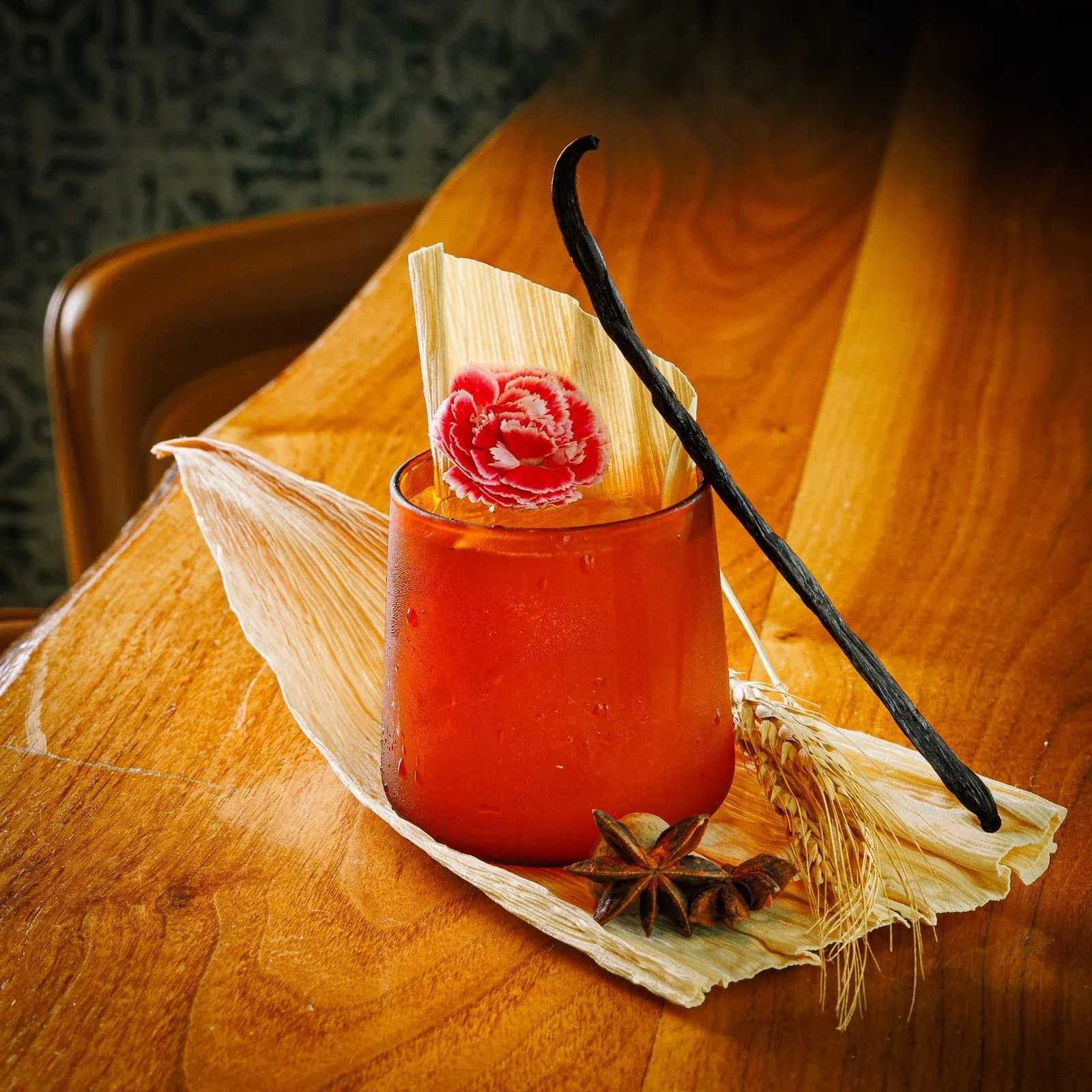 A red drink in a frosted glass, garnished with a pink carnation flower, a vanilla bean, star anise, and dried corn husks on a wooden table.