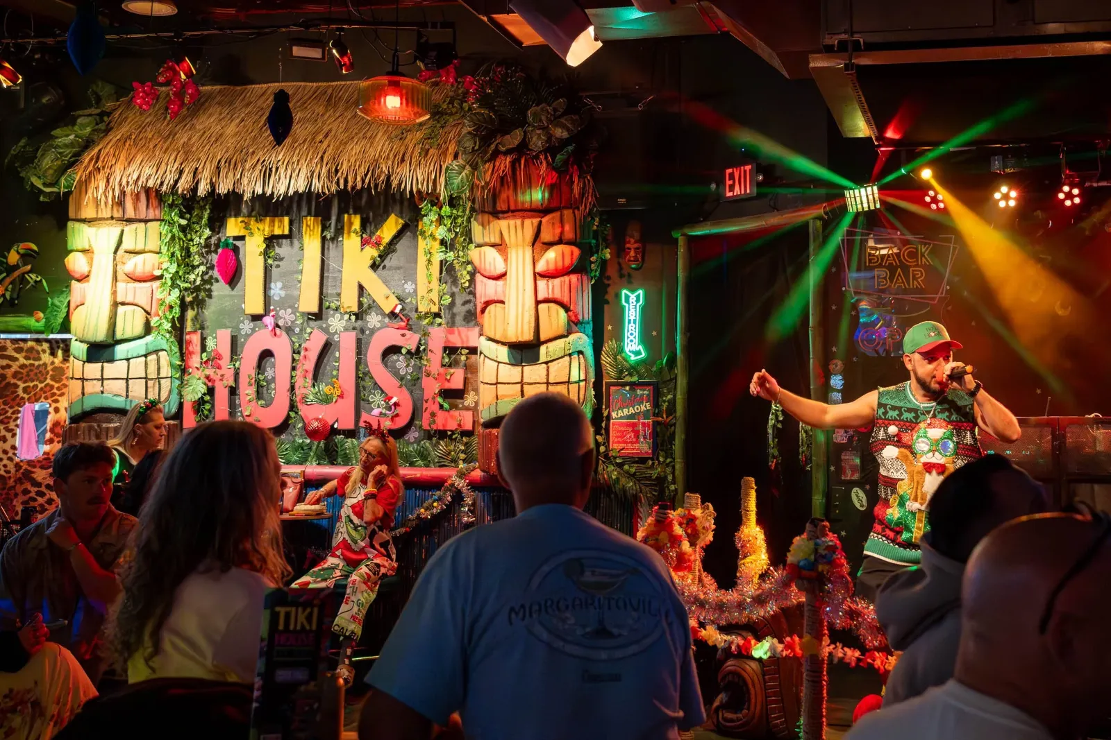 A lively tropical-themed bar scene with a colorful sign that reads 'TIKI HOUSE' decorated with tiki statues and plants. People are seated at tables, some wearing festive clothing, and a performer in a