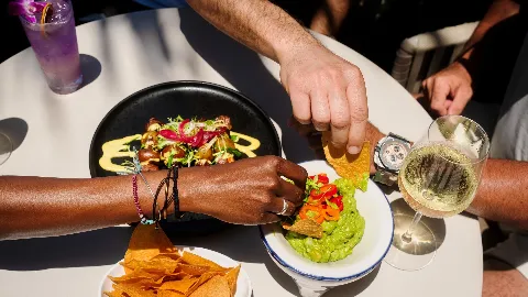 People sharing a meal at a table, including guacamole, tortilla chips, grape salad with dressing, and glasses of white wine.