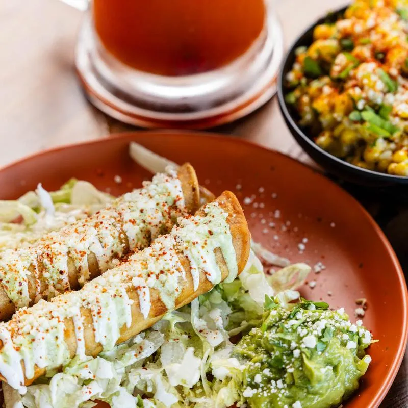 Two taquitos served with guacamole, Michelada and corn salad.