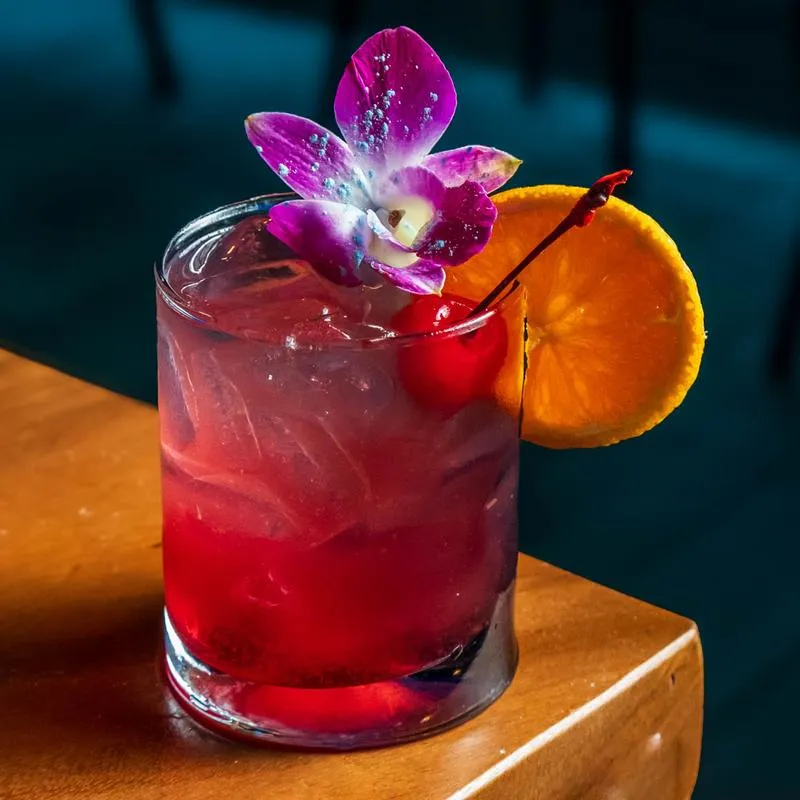 A red colored cocktail garnished with a cherry, lemon wheel, and a purple flower.