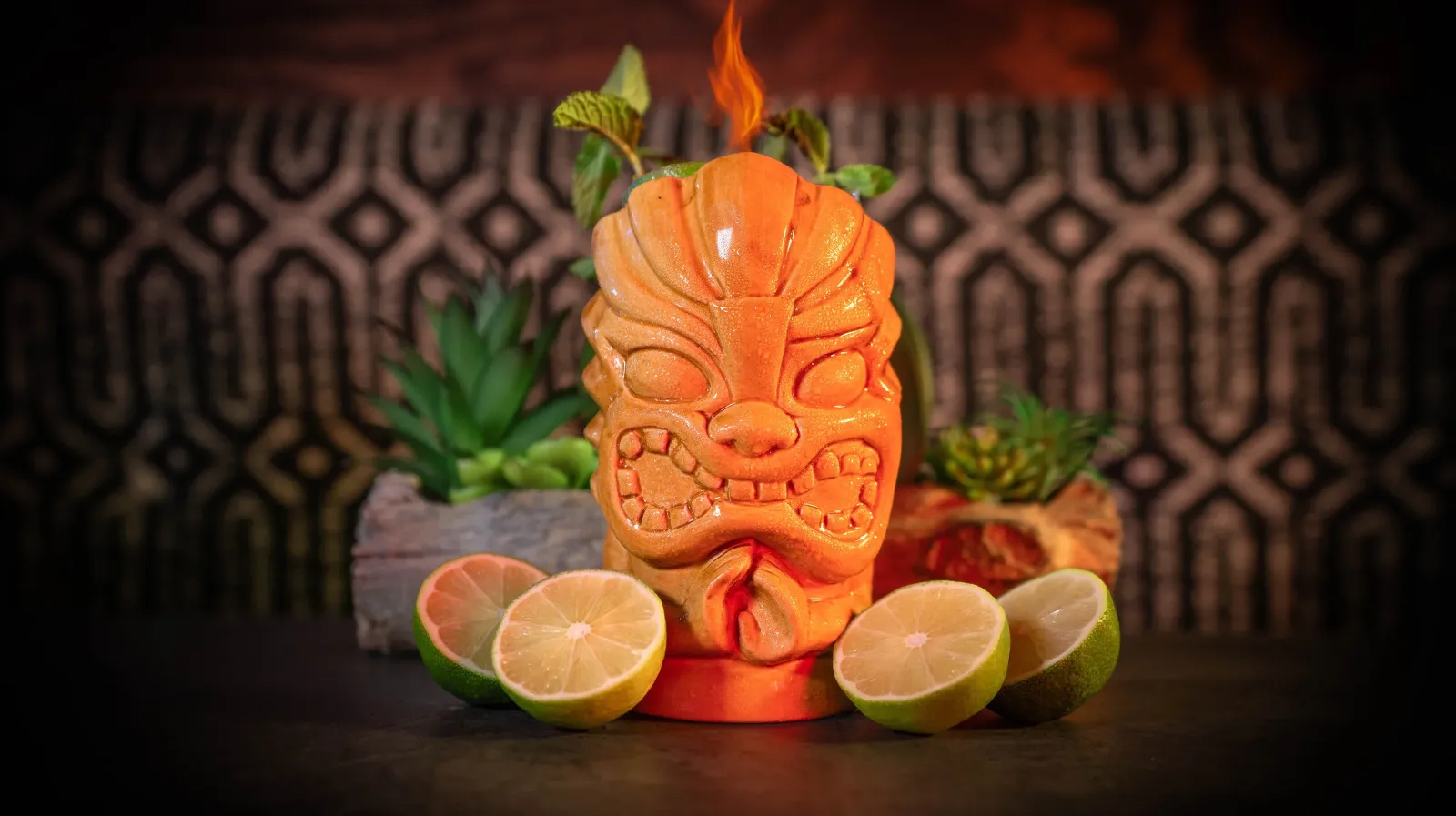 A tiki-style cocktail shaped like a tiki face with orange flame on top, surrounded by halved limes and green succulents on a dark surface with a patterned background.