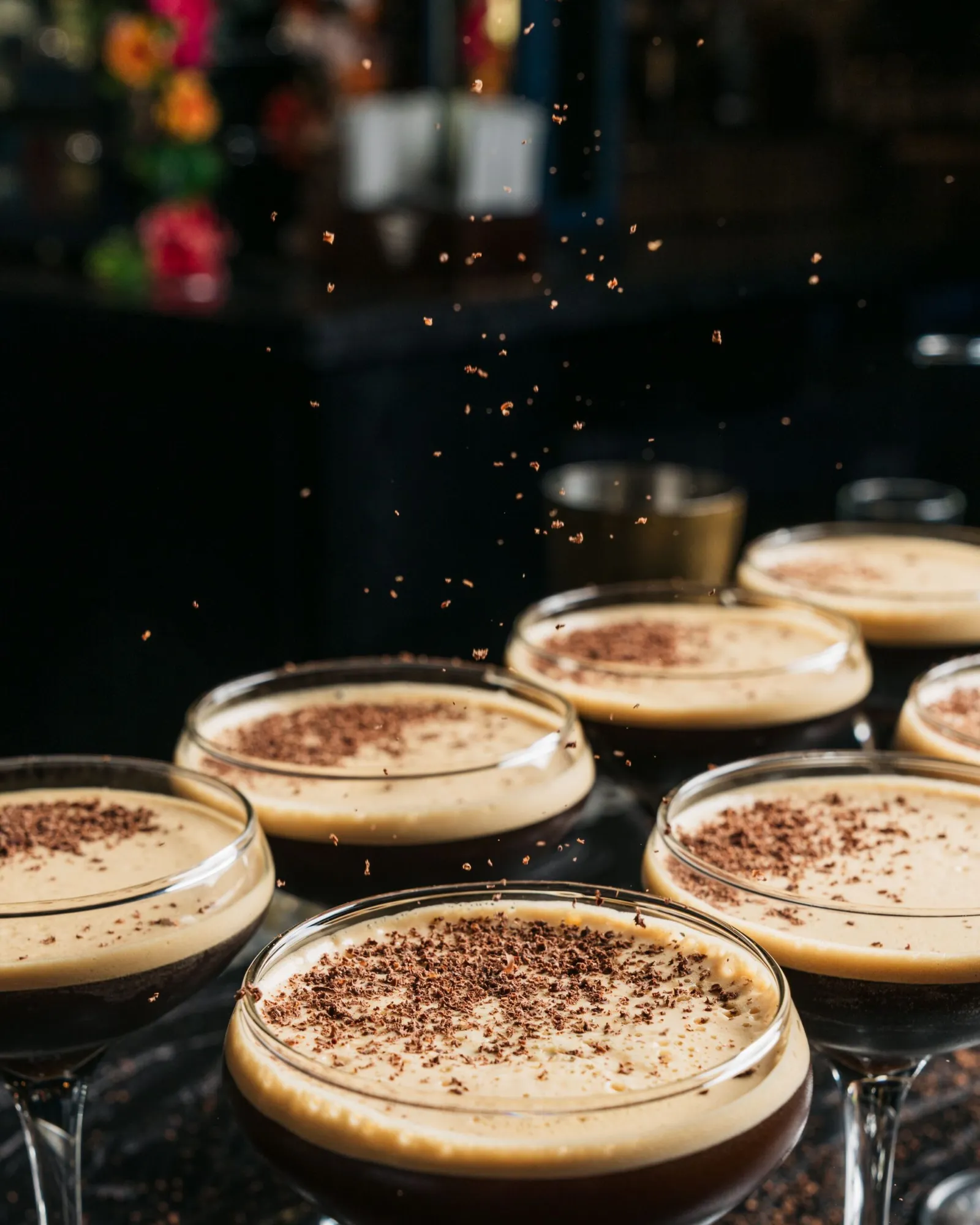 Several cocktail glasses filled with frothy espresso martinis are topped with grated chocolate, as shavings fall onto the drinks. The dimly lit, slightly out-of-focus background sets a cozy mood—perfe