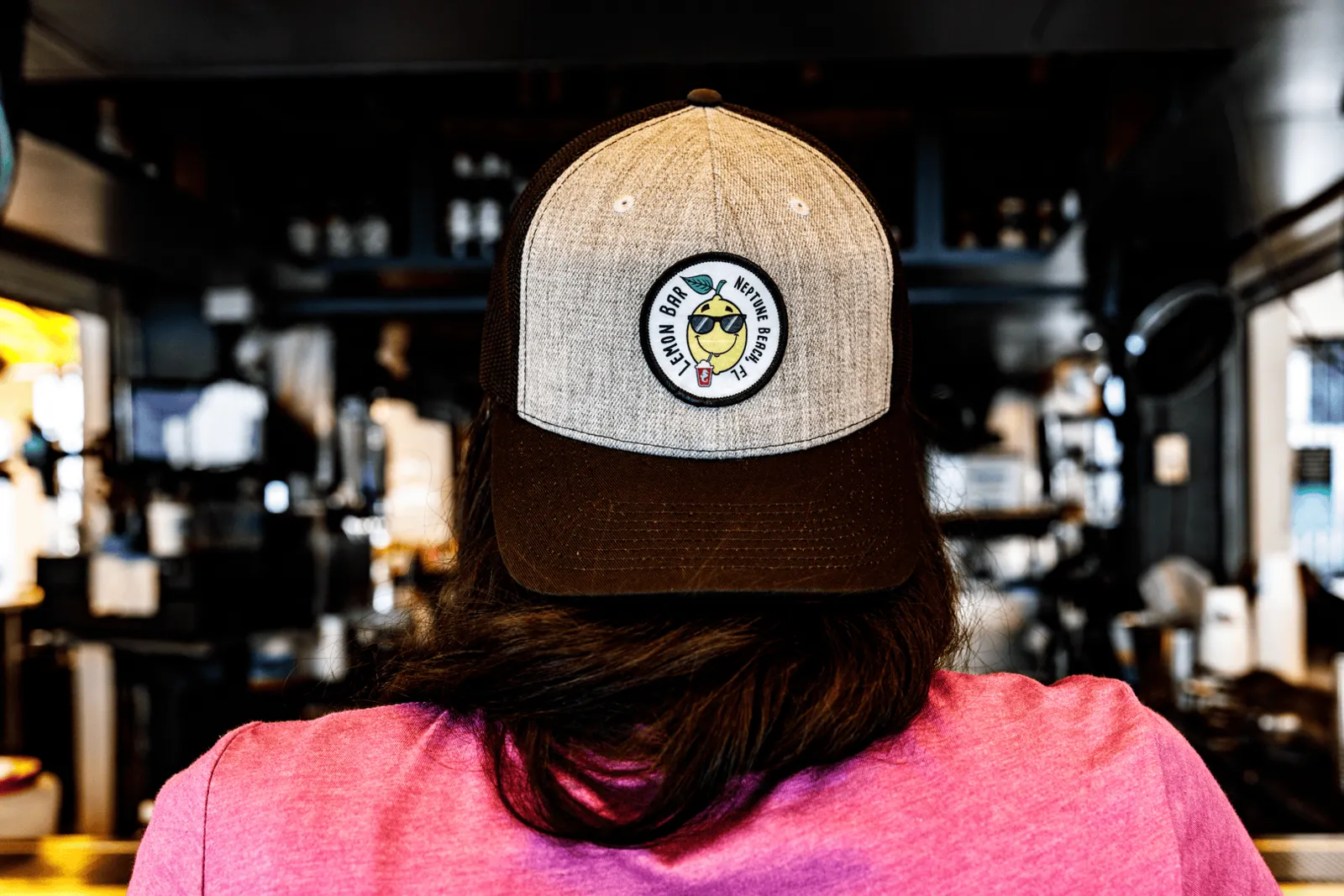 Back of a person wearing a beige and brown trucker hat with a neon bar patch, with a pink shirt inside a bar or cafe setting.