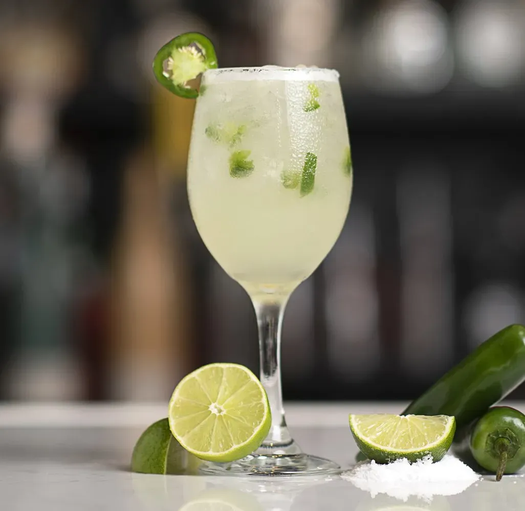A drink in a wine glass with limes and jalapenos on top.