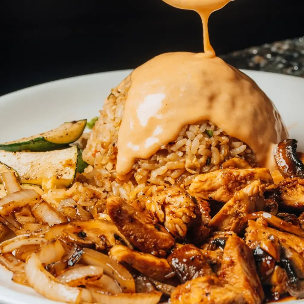 Close-up of Benihana hibachi chicken, grilled onions, and zucchini with fried rice, drizzled in signature Yum Yum sauce.