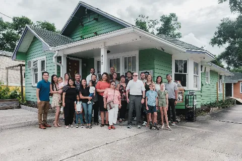 A large group of people smiling in front of a green house with the number 618 on the porch. The group includes adults and children, gathered closely for a group photo. The background features trees an