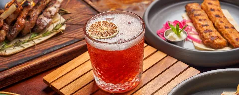 Red signature cocktail with a dried lime wheel, served beside grilled kafta skewers and flatbread