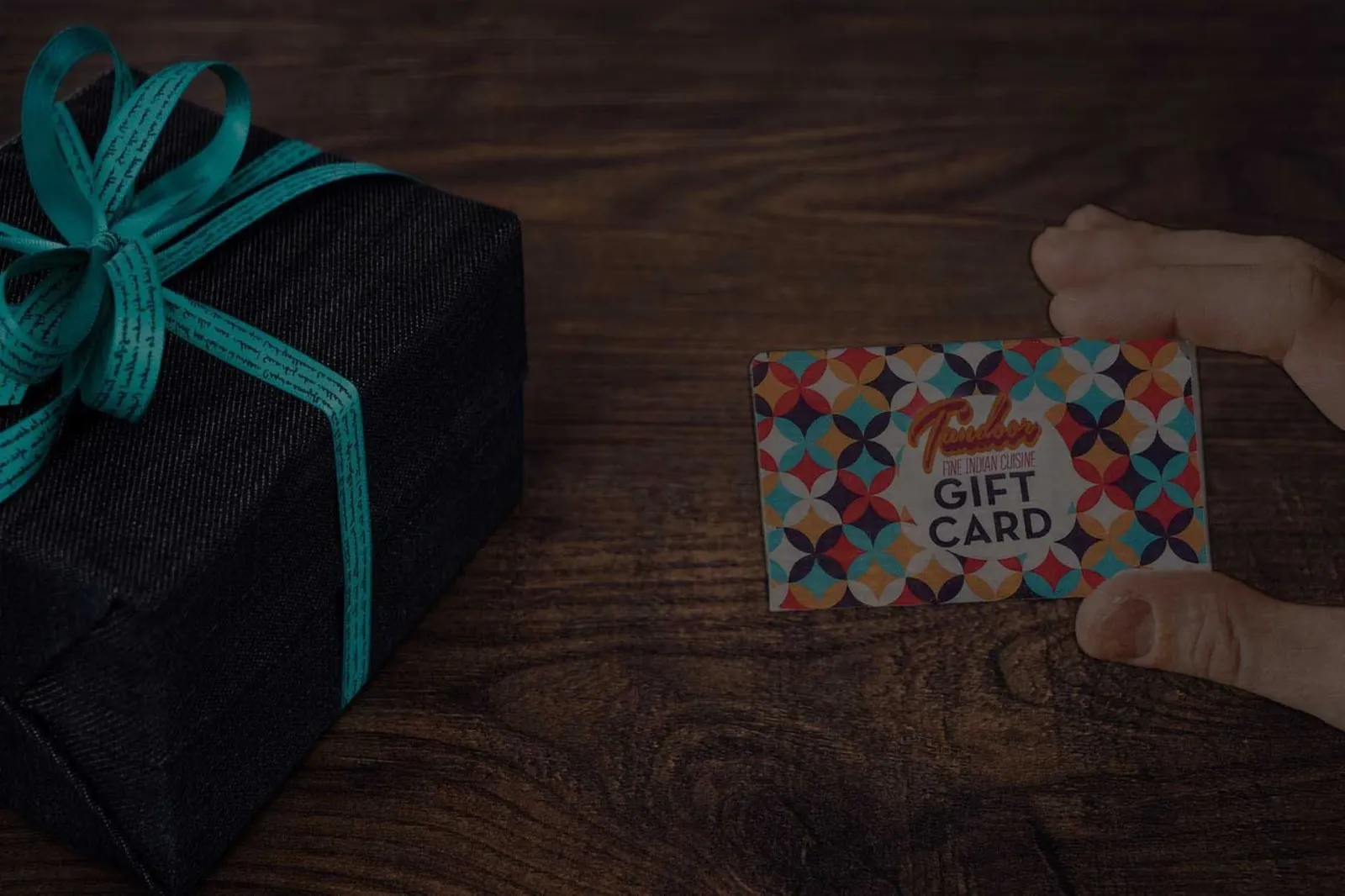 Black gift box with a teal ribbon and a colorful gift card on a wooden surface.