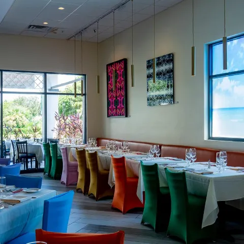 Restaurant interior with colorful chairs, white tablecloths, wall art, and large windows.