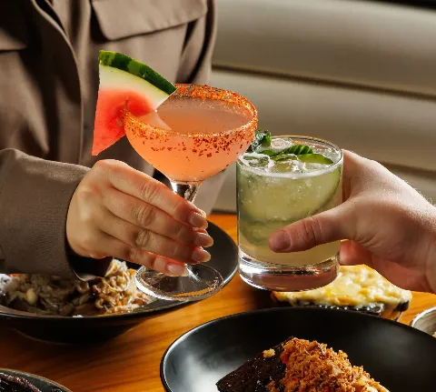 two cocktails clinking above a table spread of steak and seafood dishes