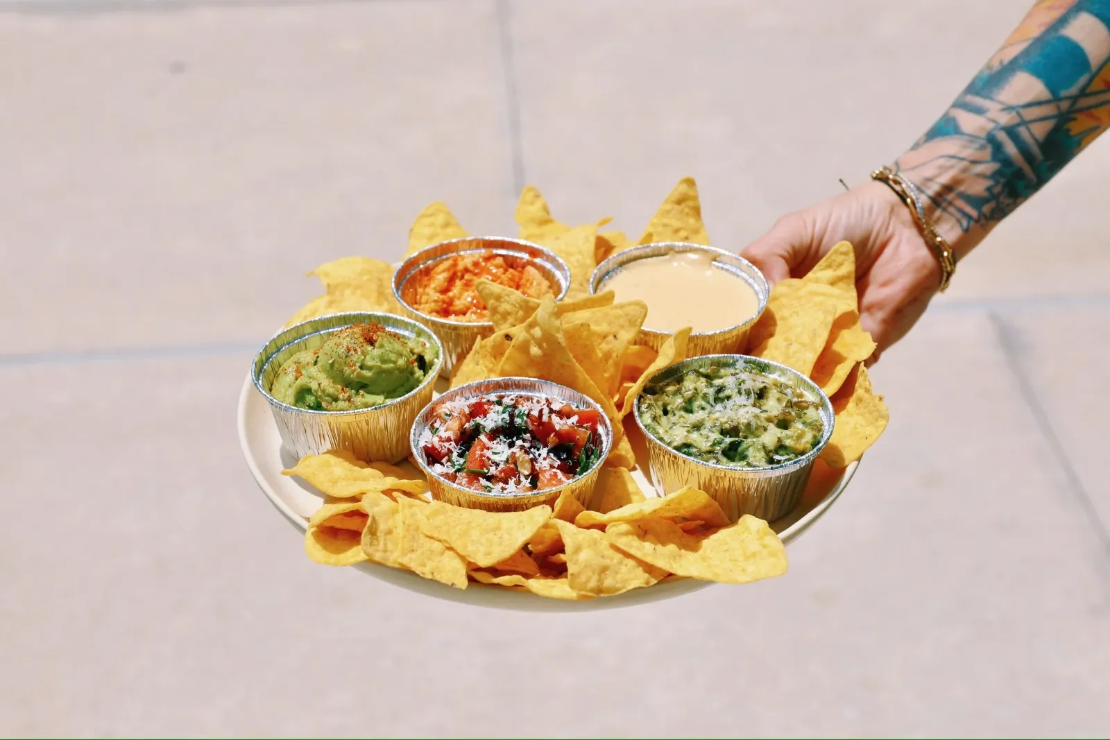 Plate with tortilla chips and five dips: guacamole, bruschetta, queso, spinach dip, and hummus, being held by tattooed hand.