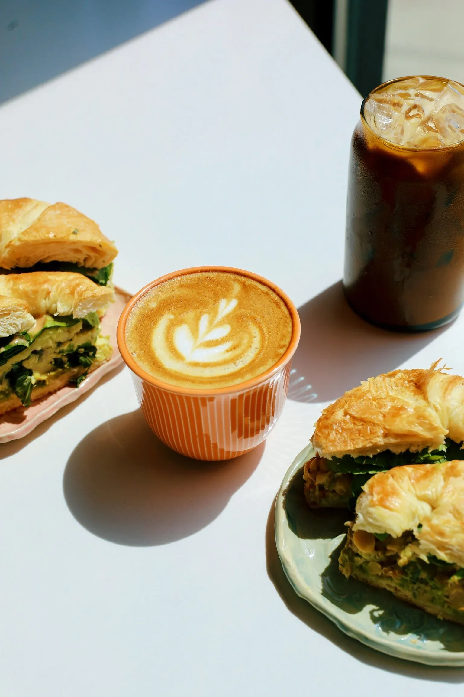 Breakfast setup with a latte, iced coffee, and croissant sandwiches on a table.