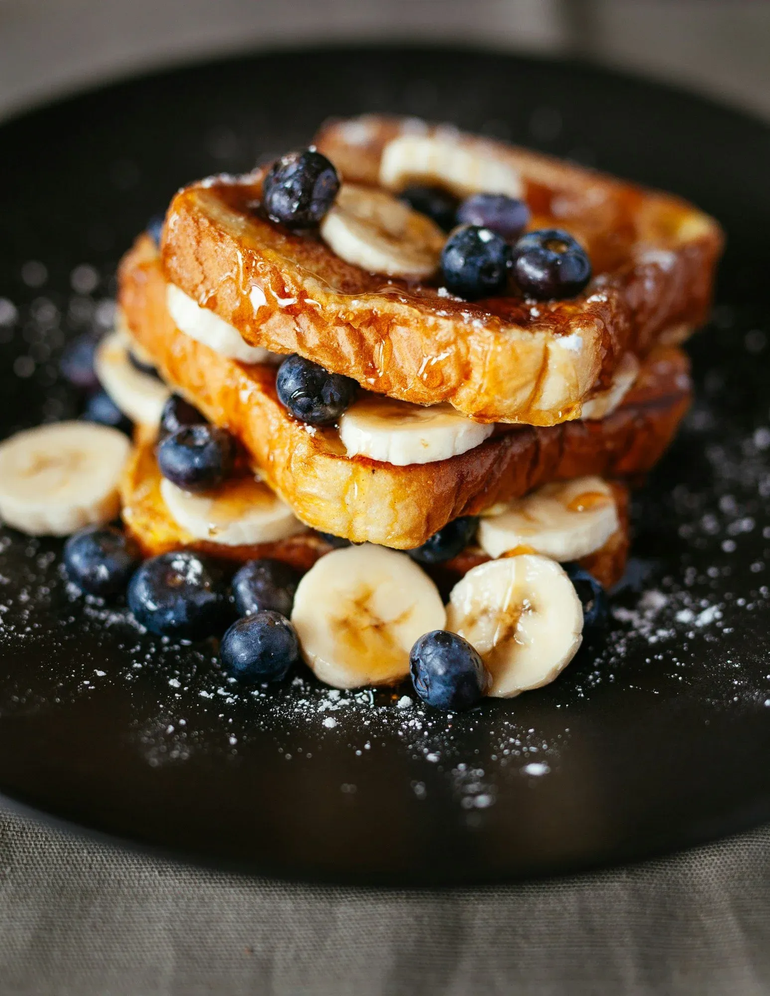 Stacked slices of French toast topped with blueberries, banana slices, and syrup, served on a black plate.