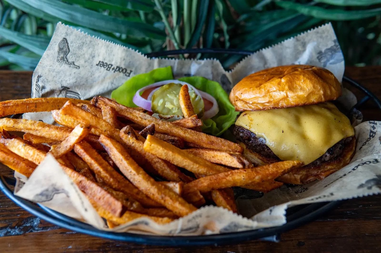 Cheeseburger with melted cheese, soft bun, lettuce, tomato, pickles, and onion slices, served with crispy French fries and a side of pickles on a basket-lined tray.