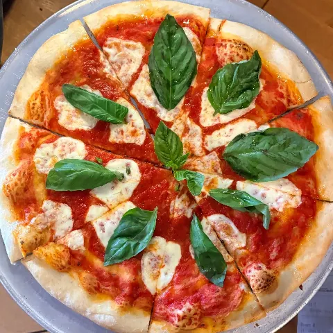 A pizza with basil leaves, tomato sauce, cheese, and a thin crust, cut into slices on a metal tray.