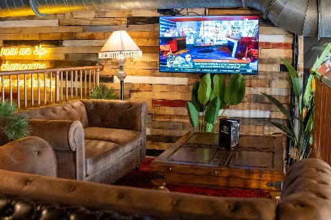 A cozy lounge area with brown leather sofas, potted plants, a wooden coffee table, a vintage floor lamp, a neon sign, and a wall-mounted TV showing a sports news broadcast in a rustic styled room with