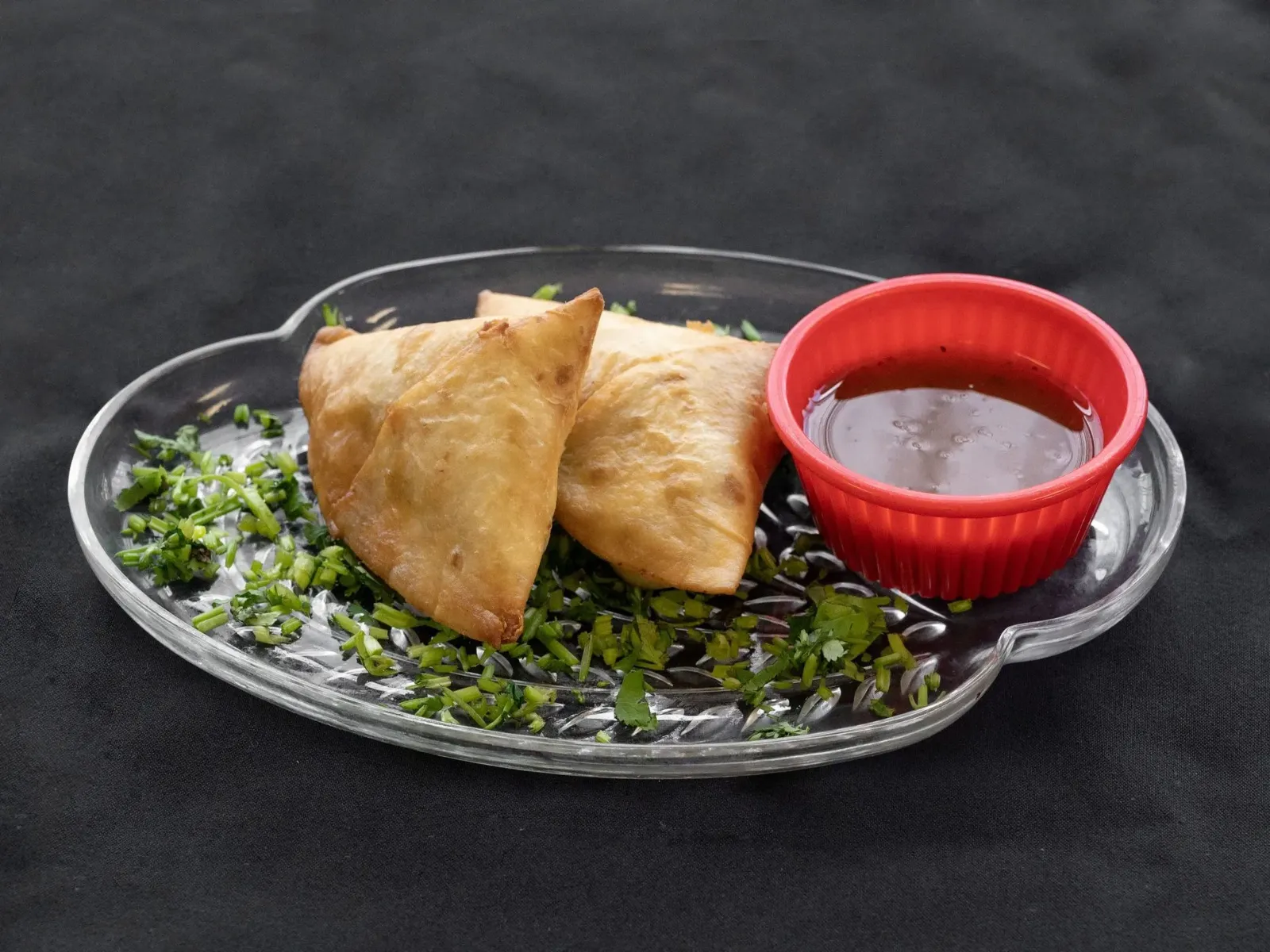 Two golden samosas with chutney on a clear plate garnished with chopped greens.
