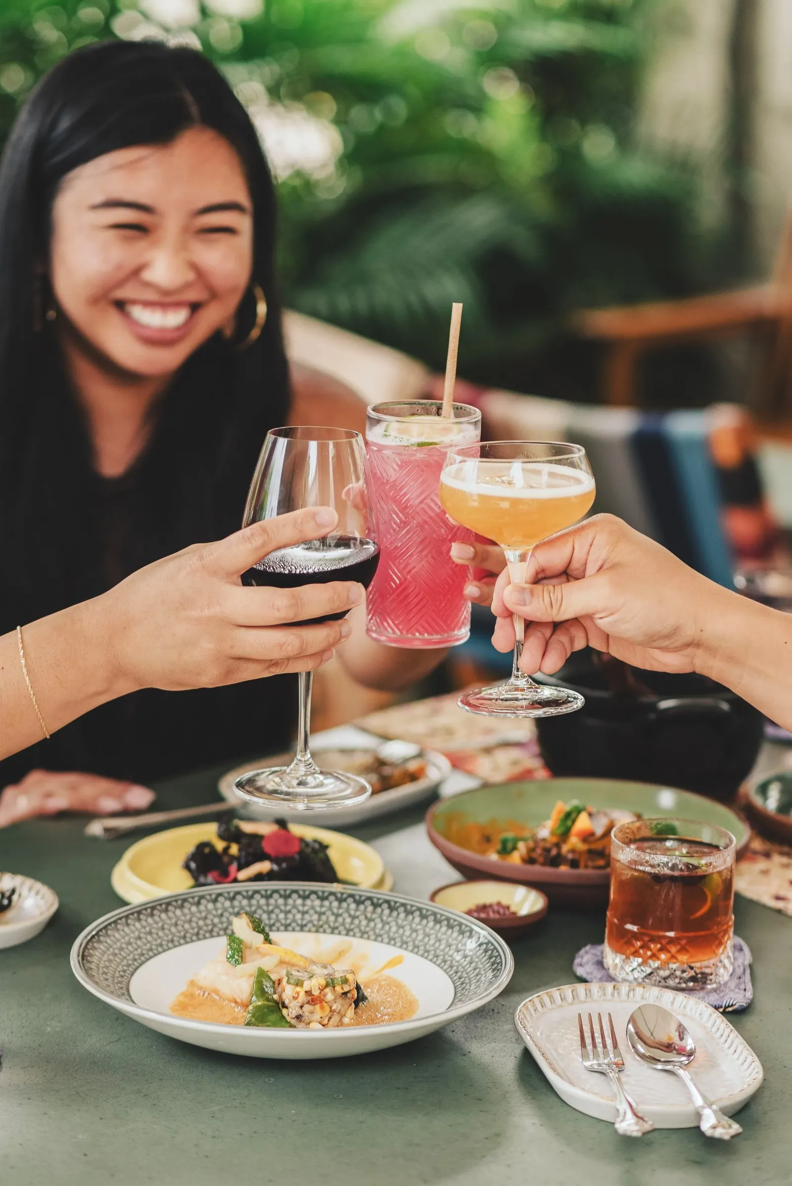 Group toasting wine glasses and cocktails over a table with various dishes, including plates with appetizers and a smiling person in the background.