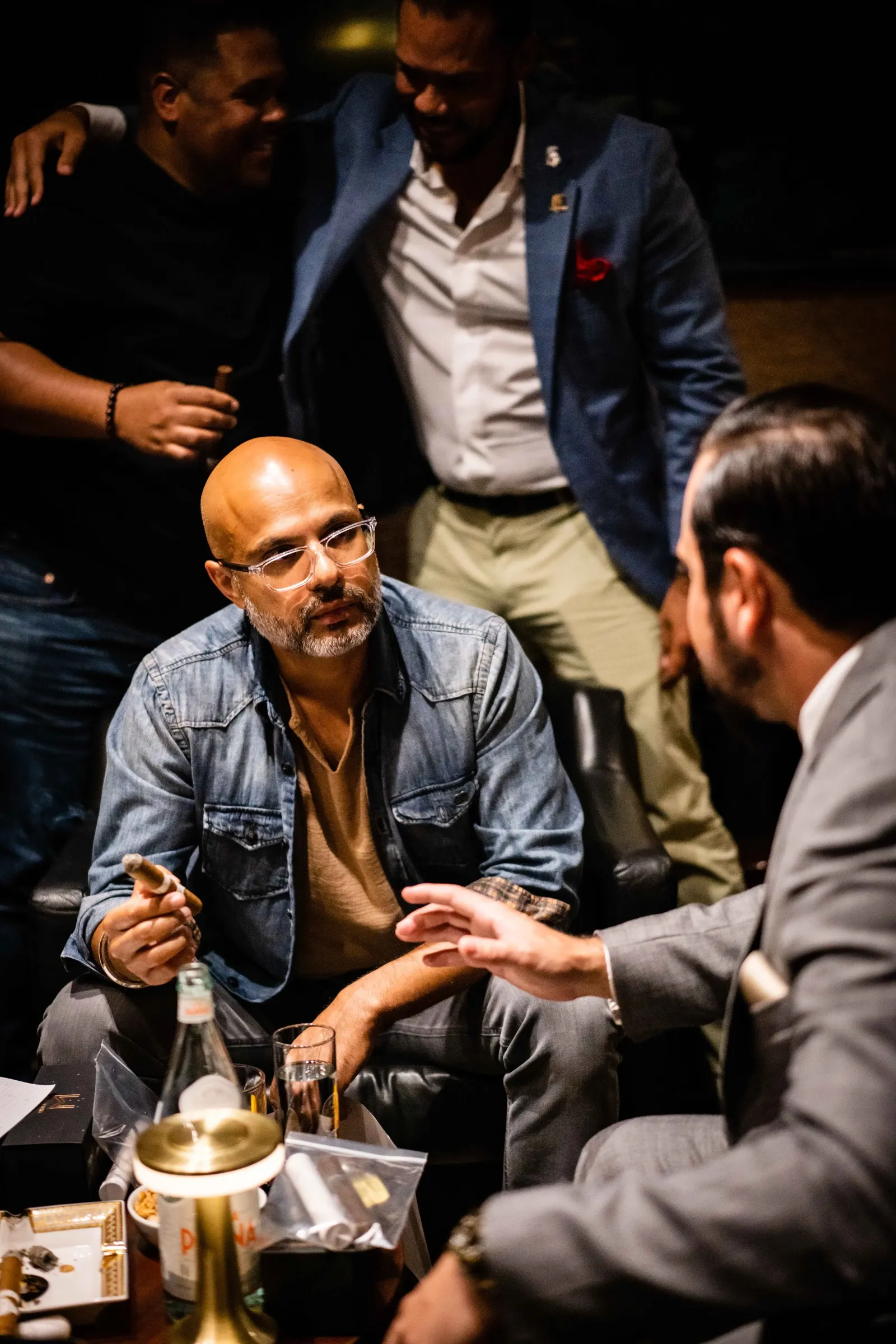 Four men in a dimly lit room are engaged in conversation. One man with glasses, a beard, and a bald head wearing a denim jacket holds a cigar and appears to be listening. Two men are leaning in toward