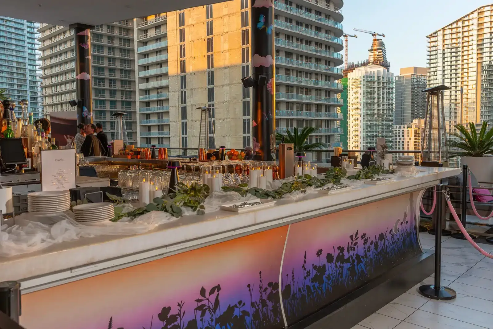 Rooftop bar with a long counter, glassware, bottles, and candles, with tall city buildings in the background.