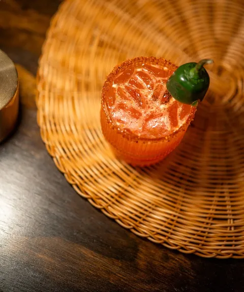 A spicy cocktail with ice and a jalapeño pepper garnish, served on a woven rattan mat.