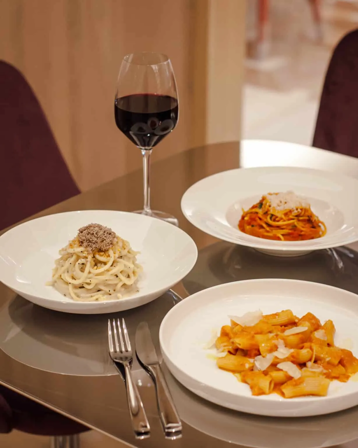 A luxurious dining table with three Italian pasta dishes including creamy truffle pasta, rigatoni, and spaghetti, paired with red wine, next to a smiling woman toasting with vibrant cocktails and a cl