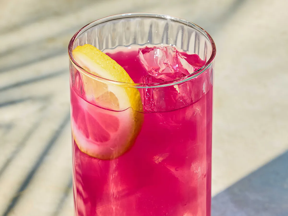 A tall glass of vibrant pink lemonade with ice, garnished with a lemon slice, sits on a sunlit surface.