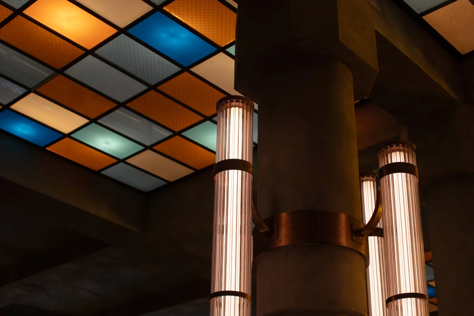 Colorful stained glass ceiling panels with warm and cool tones, green-colored vertical Edison-style light fixtures mounted on a dark wall, and a cylindrical gold-colored column.
