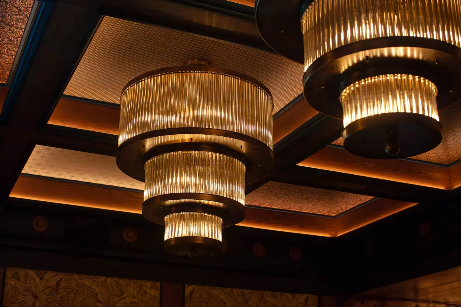 Multiple ornate bronze and gold chandeliers hanging from a ceiling with decorative patterned background, illuminated softly.