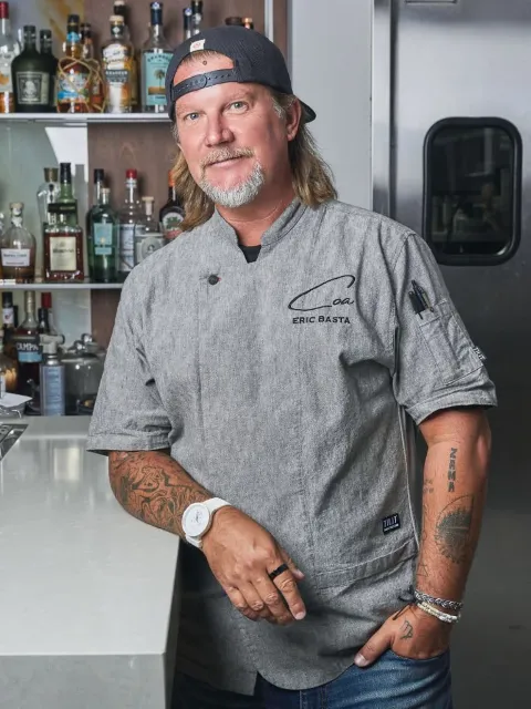 A man in a gray chef's coat with his name, Eric Basta, embroidered on it, standing in a bar or kitchen with liquor bottles on shelves behind him. He has tattoos on both arms, a white watch on his left