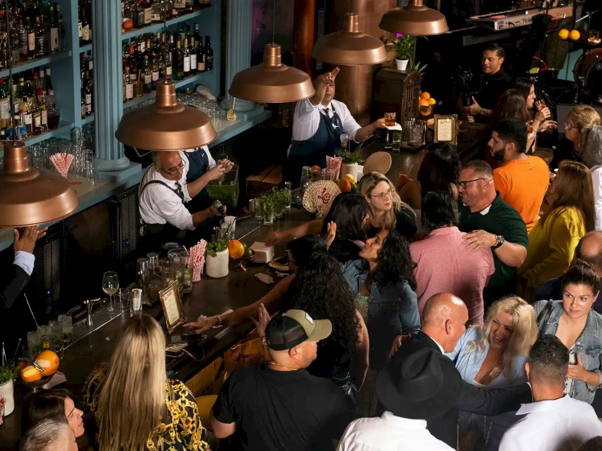 A busy bar scene with people seated at tables and a long counter, bartenders serving drinks, warm lighting, and a lively, social crowd.