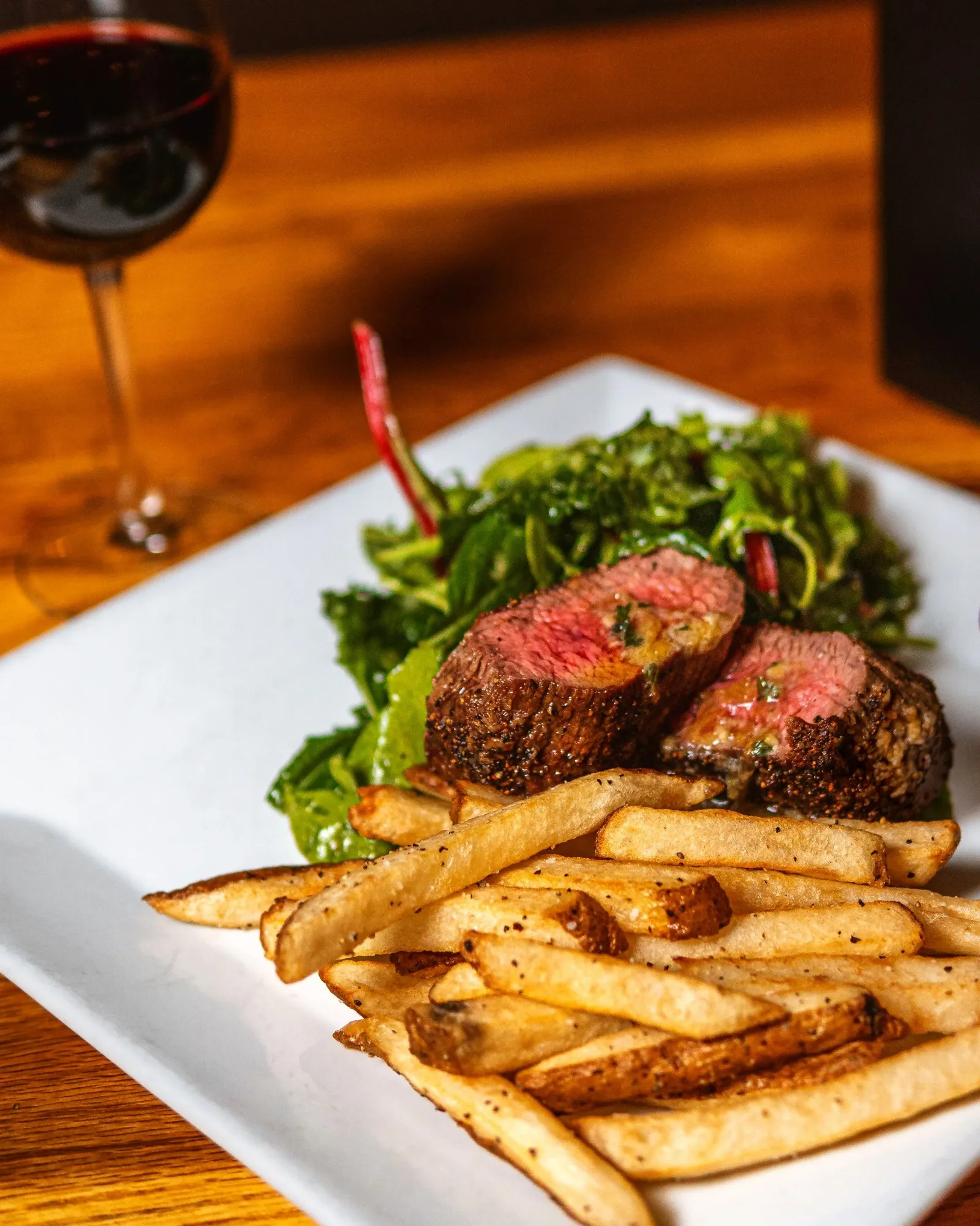 Close-up of a plate with sliced steak, french fries, and a side salad, with a glass of red wine in the background on a wooden table.