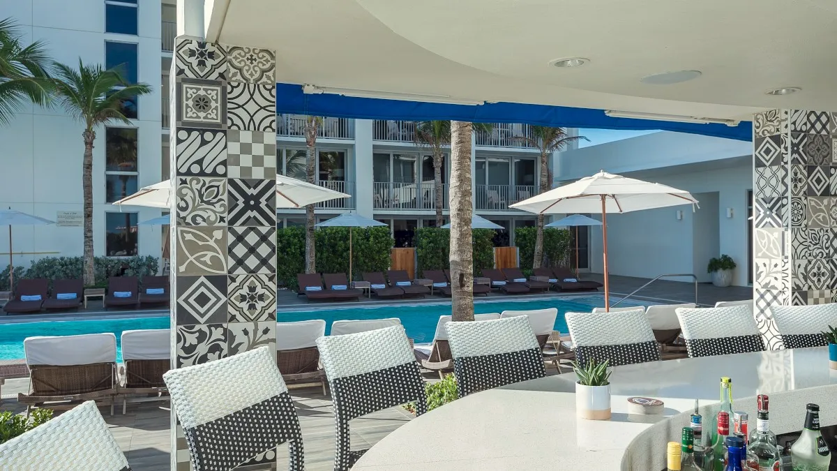 A poolside bar with patterned pillars, chairs, and a variety of bottles, set against a backdrop of lounge chairs and umbrellas.