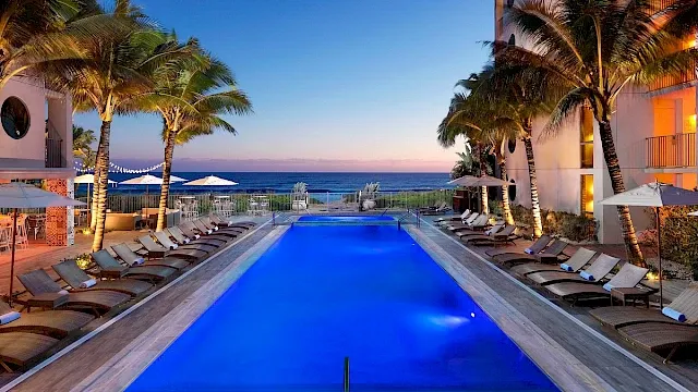 A luxurious pool lined with lounge chairs, surrounded by palm trees, overlooking the ocean at sunset, creating a serene atmosphere.