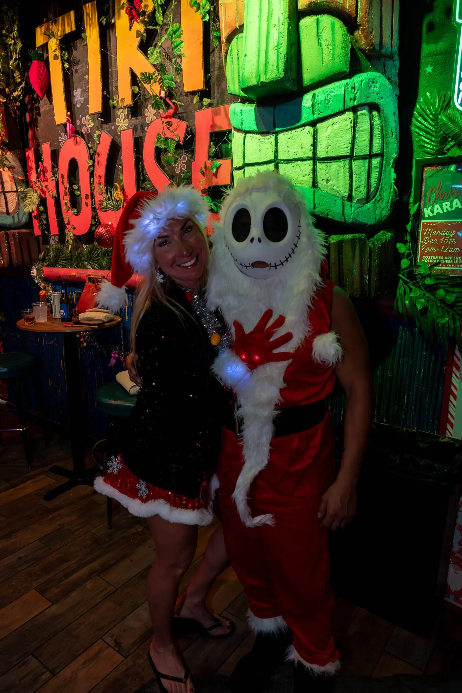 A woman and a person dressed as Jack Skellington from 'The Nightmare Before Christmas' pose together at a Christmas-themed bar decorated with colorful lights, plants, and holiday signs.