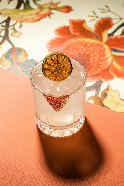 A cocktail glass with a clear drink, ice cubes, a dried lime slice garnish, and chili powder rim, on an orange surface with a floral patterned background.