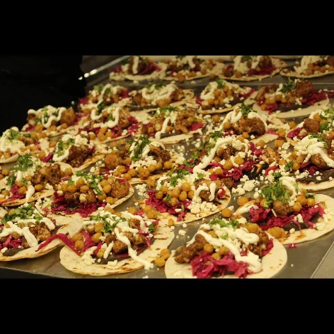 Rows of open-faced tacos on charred flatbread topped with fried chickpeas, roasted cauliflower, pickled red cabbage, crumbled feta, fresh cilantro, and a drizzle of white crema, arranged on a tray.