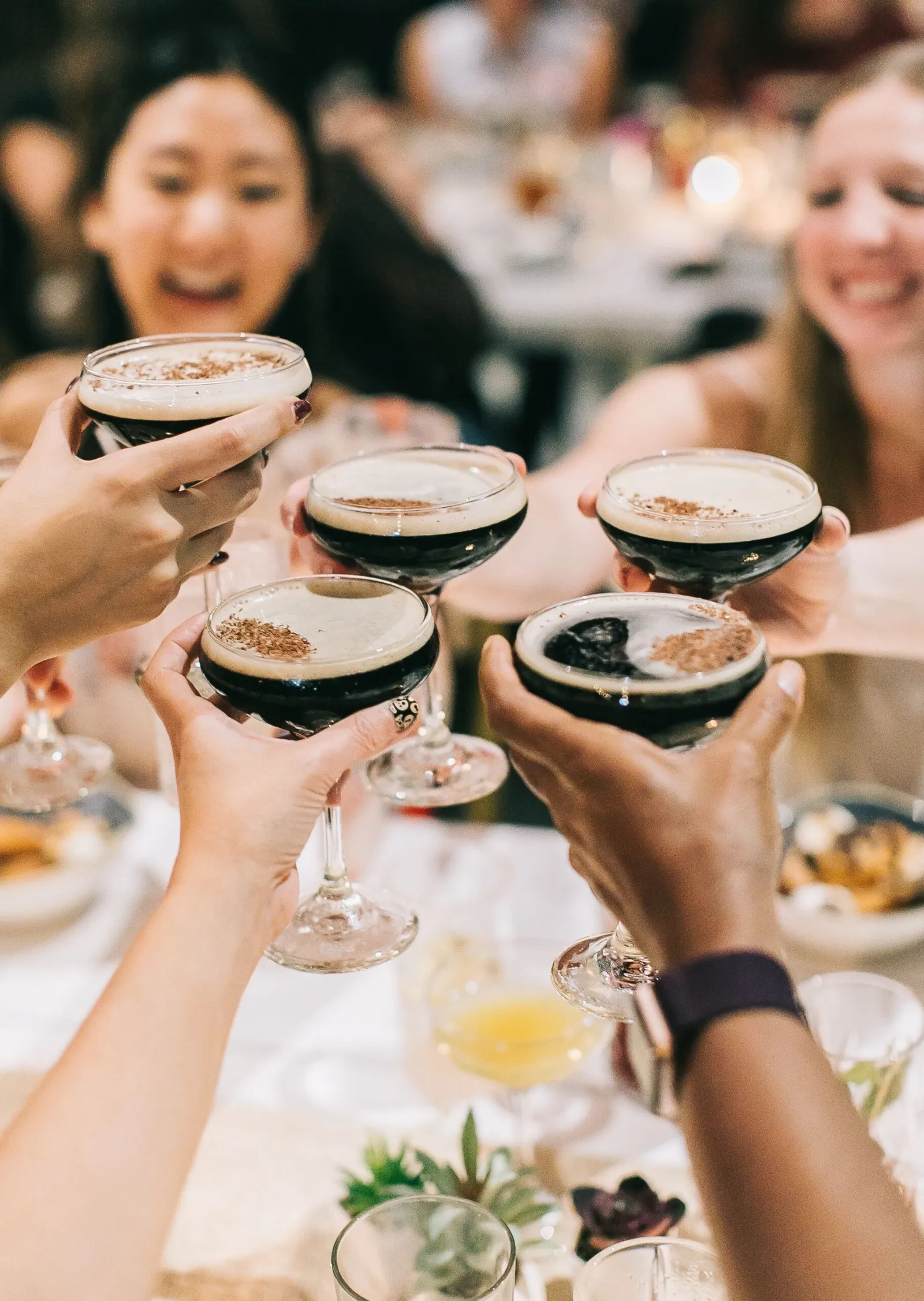 Five people clink glasses filled with a dark beverage at a lively event, smiling and celebrating around a table set with food.