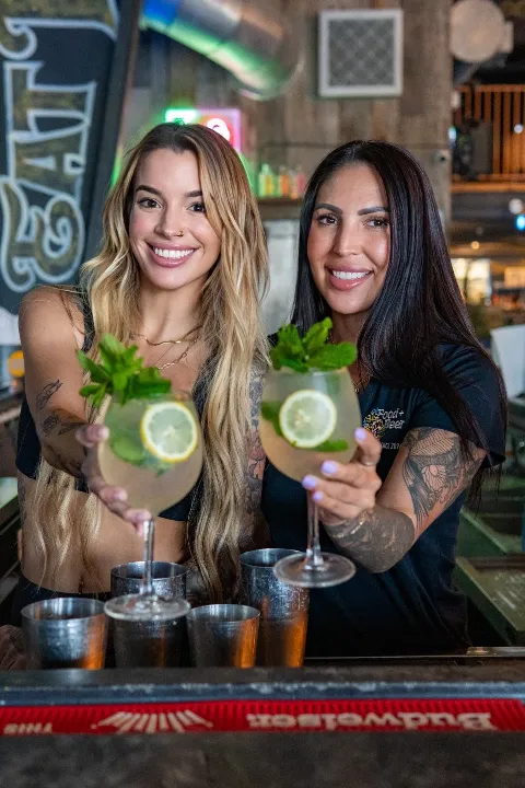 Two women smiling and holding margarita glasses with lemon slices and mint, sitting at a bar with several empty beer glasses in front of them, in a dimly lit restaurant or bar setting.