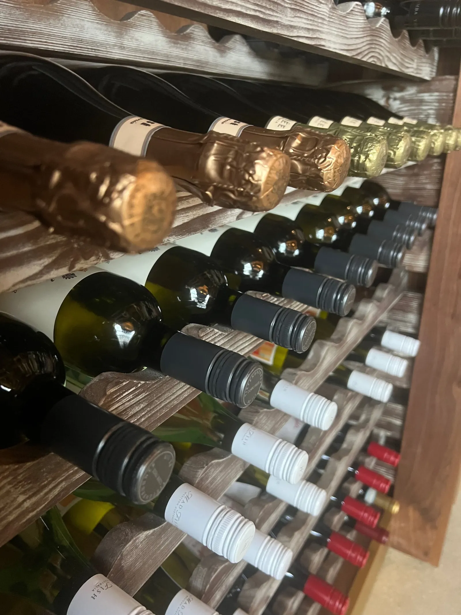 A wine rack filled with various bottles of wine, including sparkling wine with foil tops, and bottles with screw tops. The bottles are organized on wooden shelves, displayed horizontally.