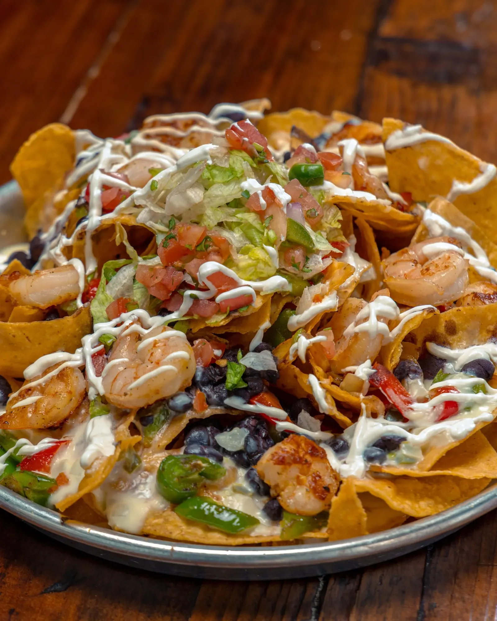 A plate of nachos with shrimp, black beans, shredded cheese, diced tomatoes, jalapeños, shredded lettuce, sour cream, and salsa on a wooden table.