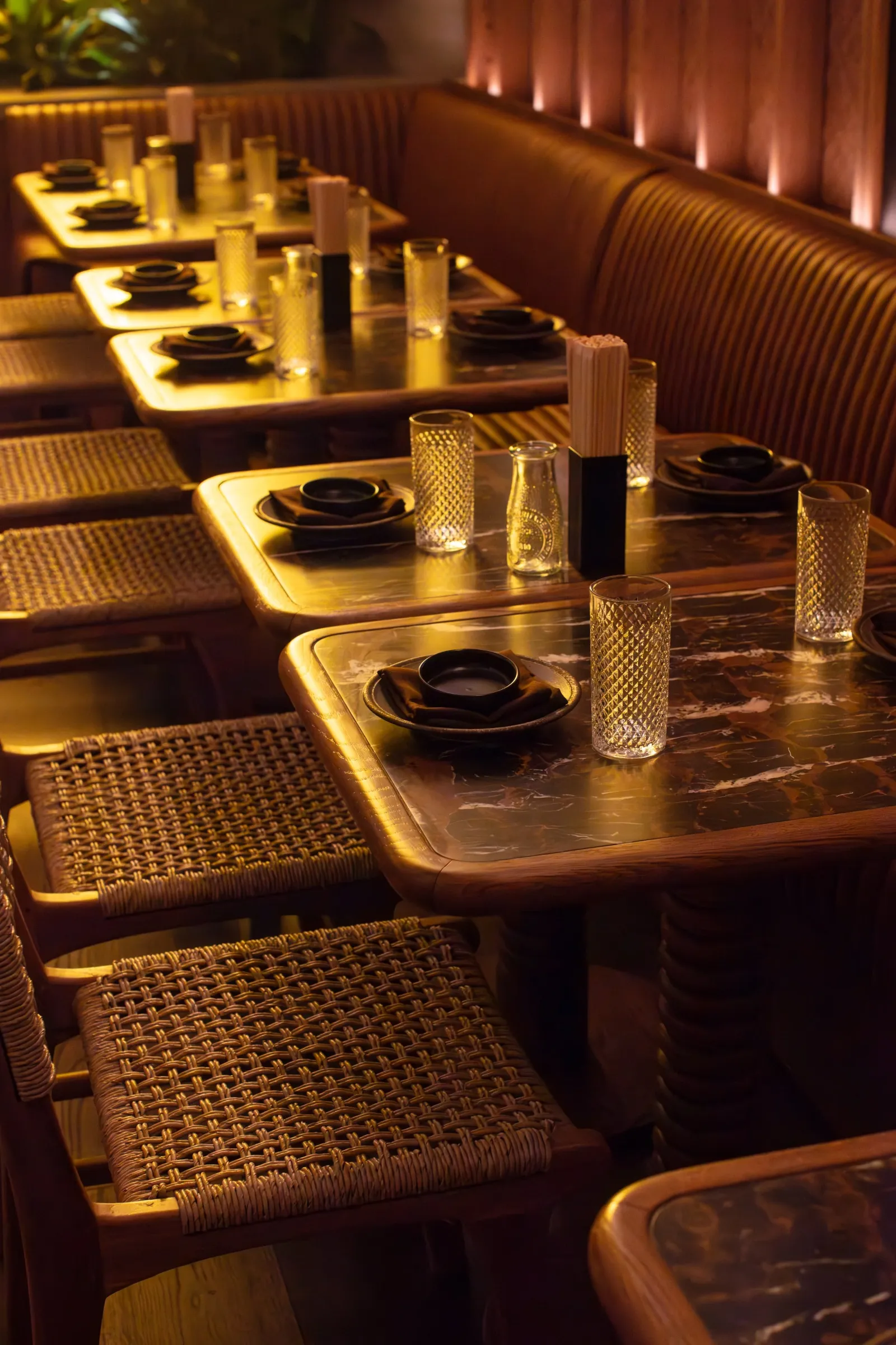 Interior of a dimly lit restaurant with several tables set for dining, featuring black plates, glasses, napkins, and chopsticks, with woven rattan chairs and a wooden-paneled wall.