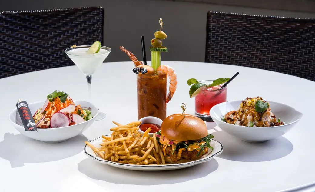 Table with drinks and food, including a burger, fries, salads, and cocktails, in an outdoor setting.