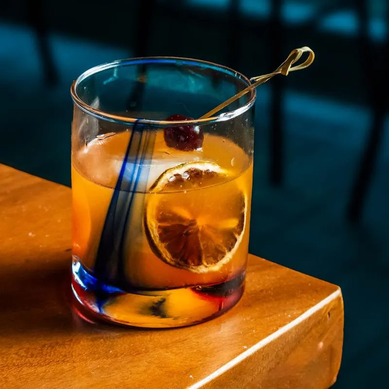 A glass of Old Fashioned resting on the corner of a table inside.
