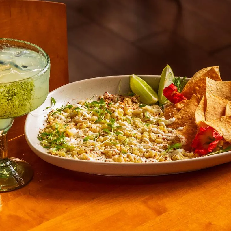 Creamy esquites with lime wedges and chips, accompanied with a margarita cocktail.